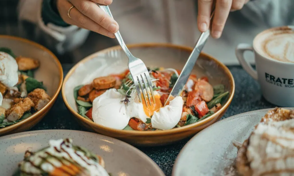 A person cuts into a poached egg on a salad with a fork and knife. Other plates of food and a cup of coffee are visible on the table.