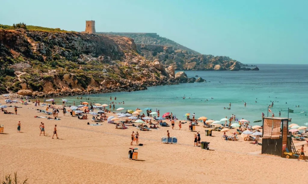 Crowded sandy beach with people sunbathing and swimming, umbrellas scattered across the shore, and rocky cliffs with a stone tower in the background.