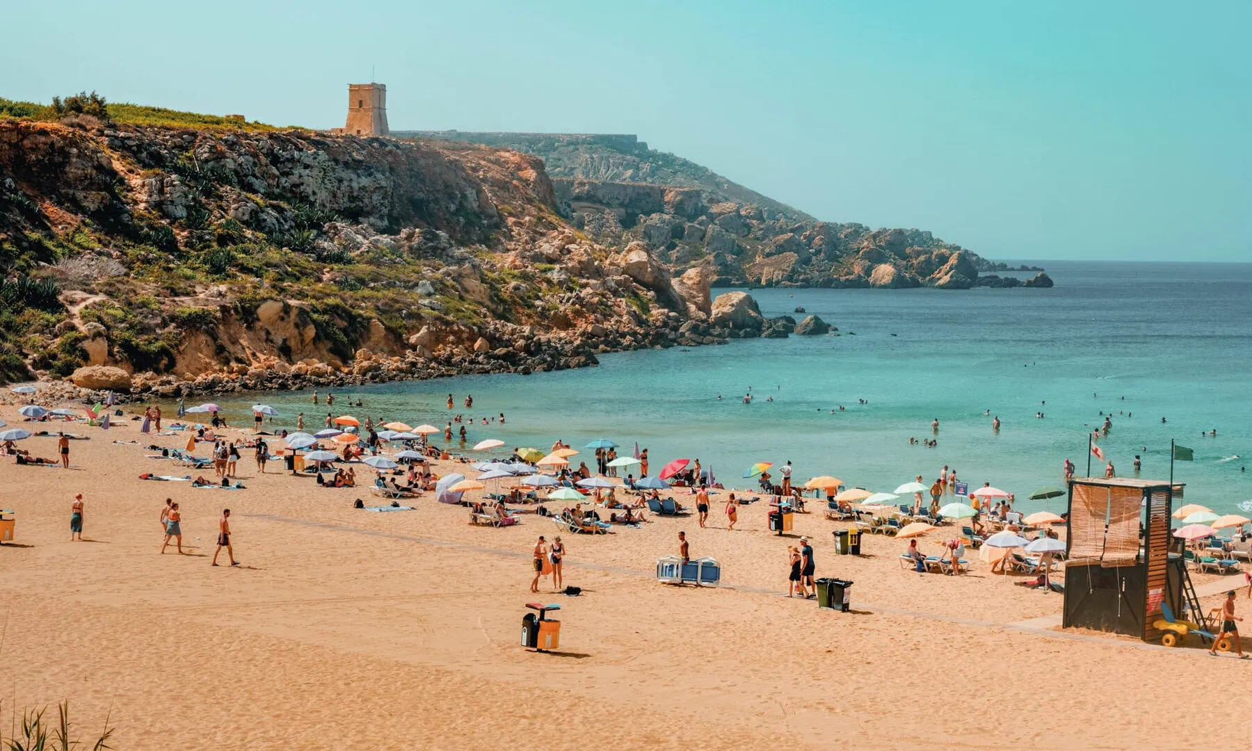 Crowded sandy beach with people sunbathing and swimming, umbrellas scattered across the shore, and rocky cliffs with a stone tower in the background.