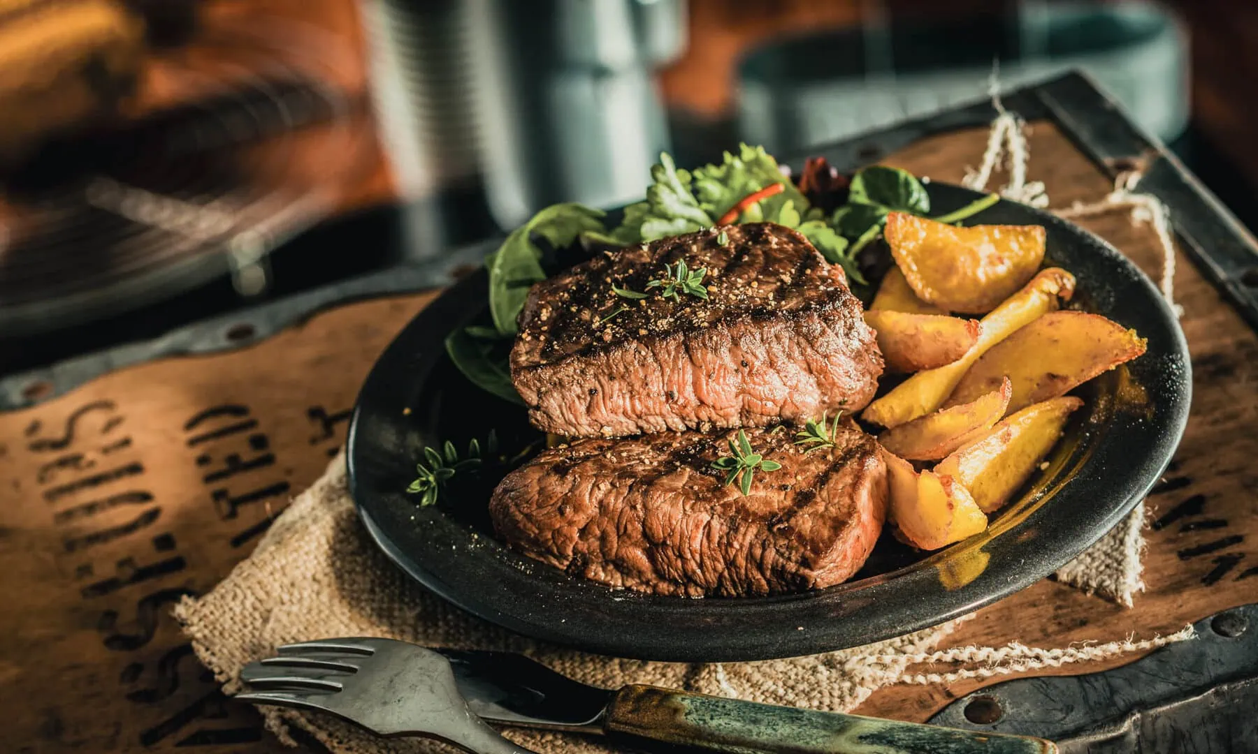 A plate with two grilled steaks, roasted potato wedges, and mixed greens on a rustic wooden table setting.