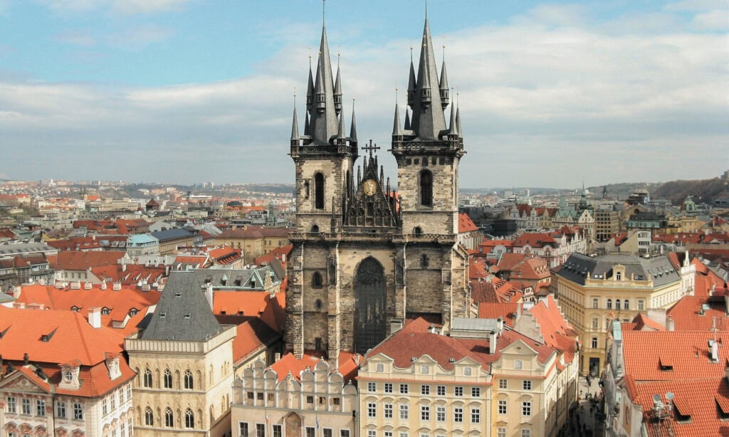 Aerial view of the Church of Our Lady before Týn with two tall Gothic spires, surrounded by historic buildings with red rooftops in Prague's Old Town.