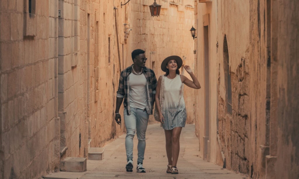 Two people walk side by side down a narrow, sunlit stone alleyway. One wears a hat and a dress; the other wears a plaid shirt, white t-shirt, and jeans.