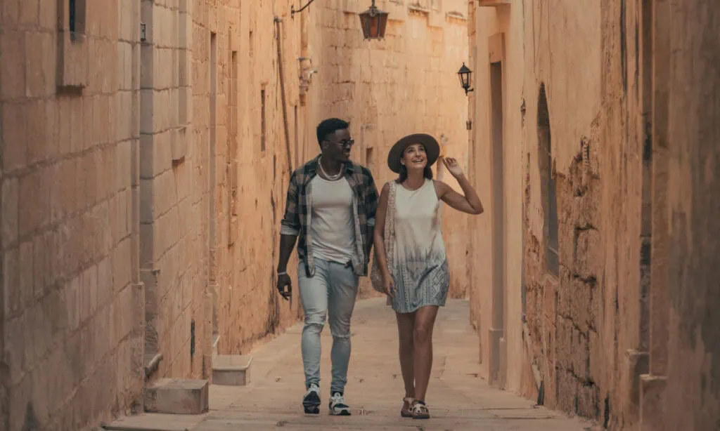 Two people walk side by side down a narrow, sunlit stone alleyway. One wears a hat and a dress; the other wears a plaid shirt, white t-shirt, and jeans.