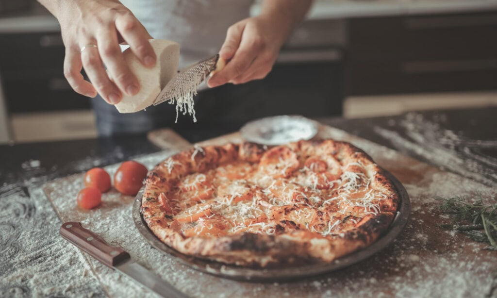 A person grates cheese onto a freshly baked pizza on a floured countertop, with tomatoes, a knife, and herbs nearby.