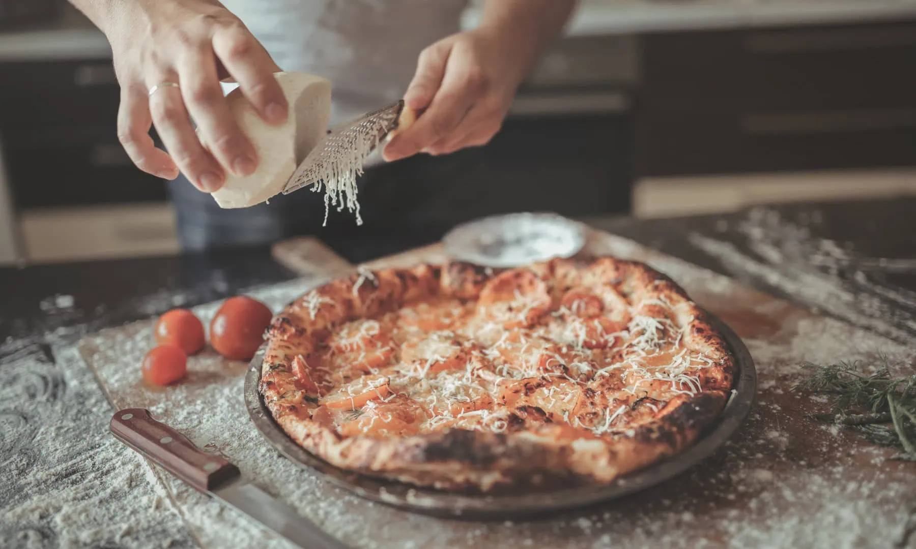 A person grates cheese onto a freshly baked pizza on a floured countertop, with tomatoes, a knife, and herbs nearby.