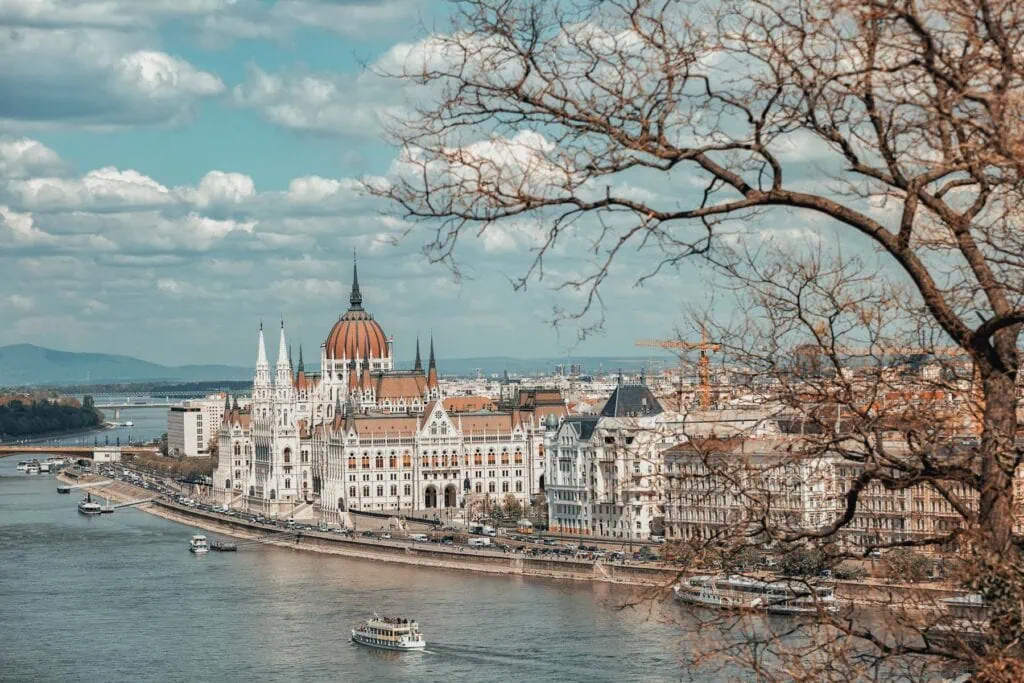 A large neo-Gothic building with a red dome sits beside a river, boats on the water, and bare tree branches in the foreground under a partly cloudy sky.