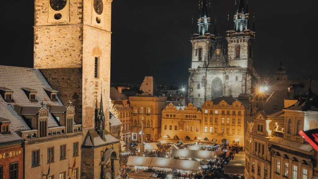 Nighttime view of a bustling Christmas market in a historic European square, with illuminated gothic church towers and old buildings surrounding the festive market stalls.