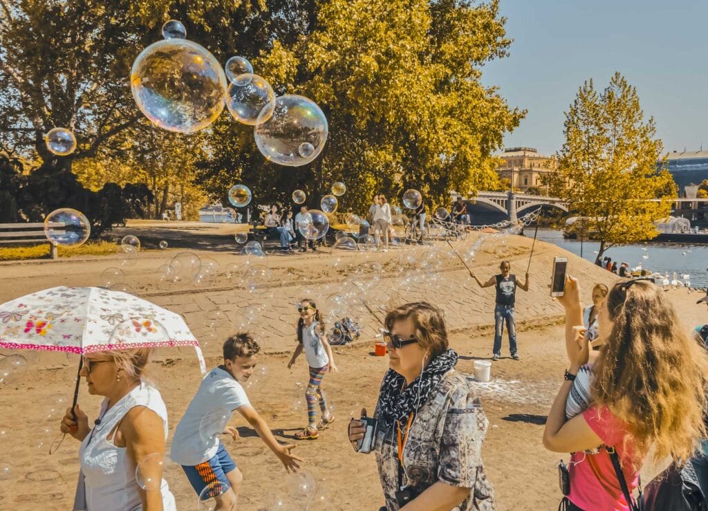 People enjoying a sunny day by a river, with large soap bubbles floating in the air and trees in the background. Some people are taking photos and children are playing.