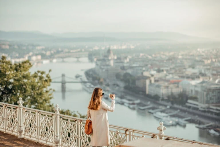 A person in a light coat takes a photo of a cityscape and river from an elevated viewpoint, with bridges and buildings visible in the distance.