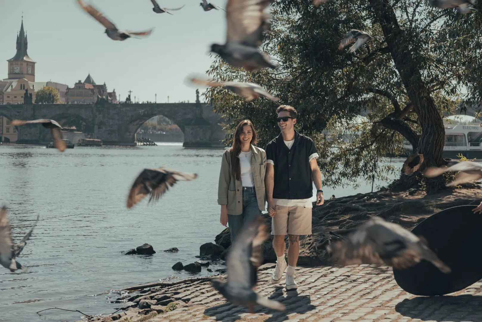 A young couple walks along a riverbank with pigeons flying nearby; a stone bridge and historic buildings are visible in the background.