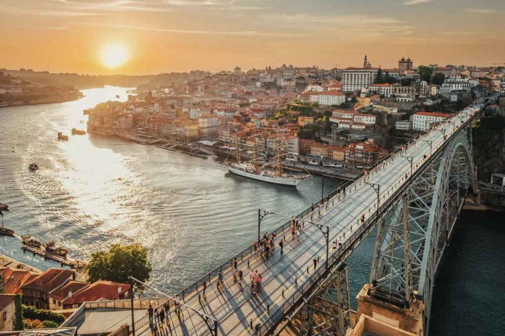 Sunset view of a cityscape with a large bridge spanning a river, pedestrians on the bridge, and a tall ship sailing below.