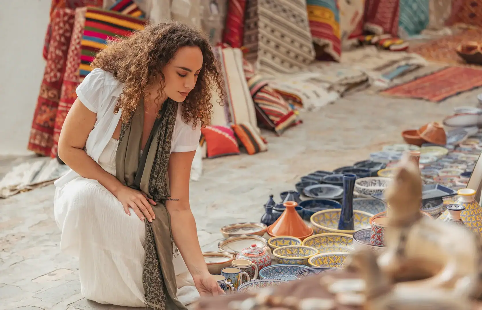 A woman in a white dress examines colorful ceramic bowls and plates displayed on the ground at a market with patterned textiles in the background.