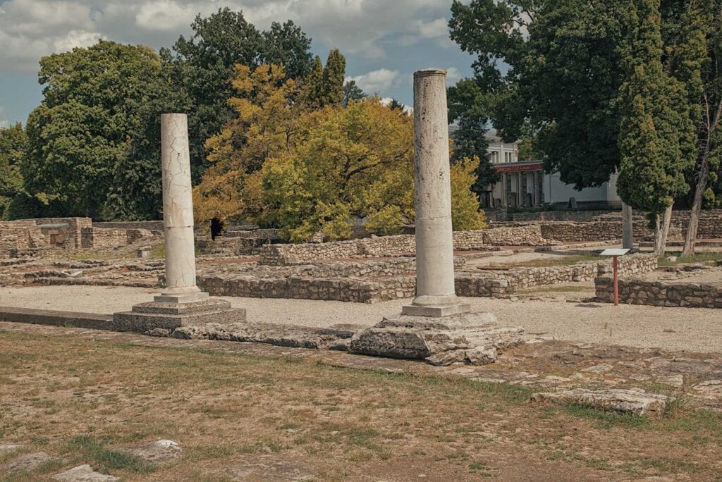 Two ancient stone columns stand among the ruins of an archaeological site, surrounded by trees and stone walls under a partly cloudy sky.