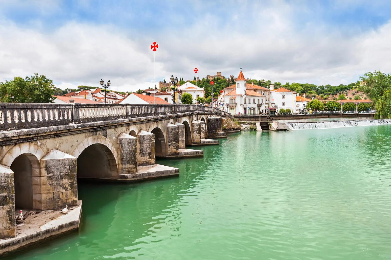 Stone arch bridge over a turquoise river in a small town with white buildings, red roofs, and flags featuring red crosses; greenery and cloudy sky in the background.