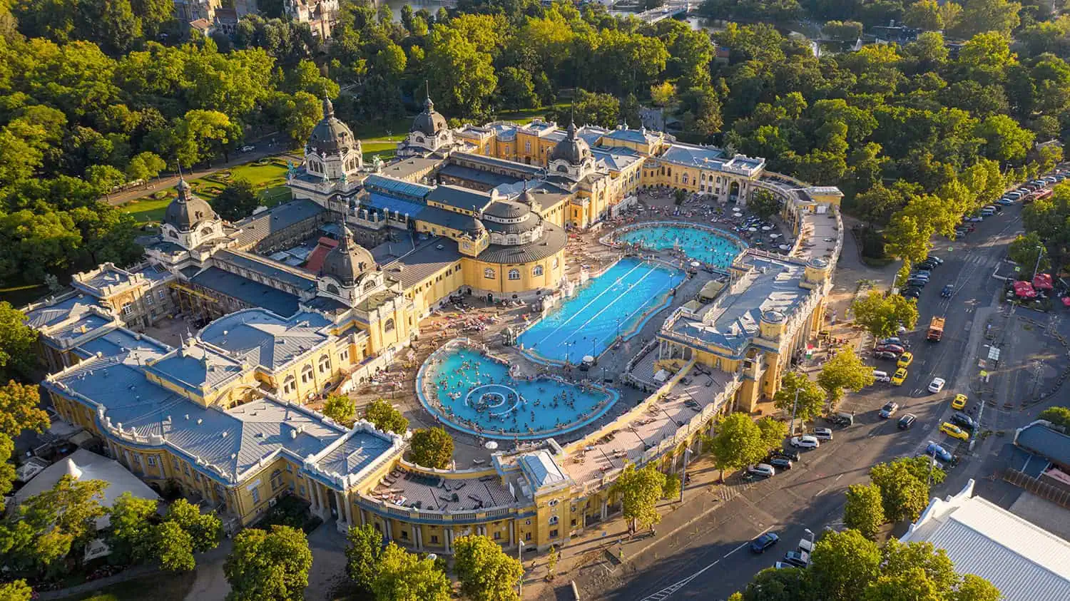 Aerial view of a large, historic yellow building complex with multiple outdoor swimming pools, surrounded by greenery and roads.