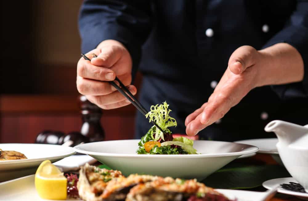 Chef meticulously plating a dish with greens and garnish, using tweezers for precision. Several other plated dishes are partially visible in the foreground.