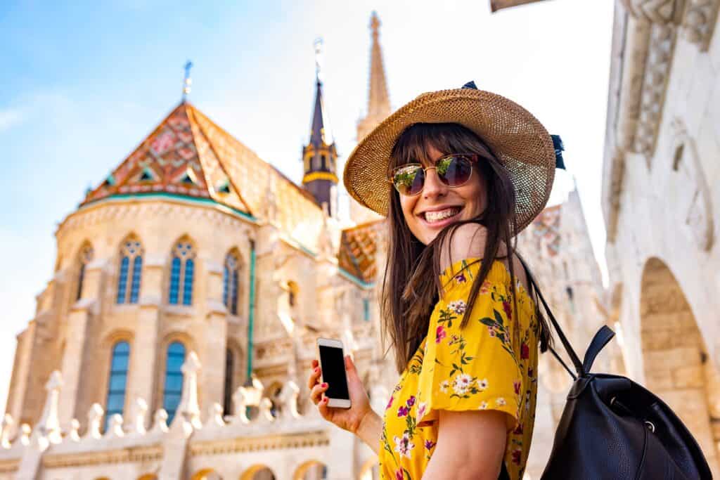 A woman in a yellow floral dress and sunhat smiles while holding a phone in front of a historic building with ornate architecture.