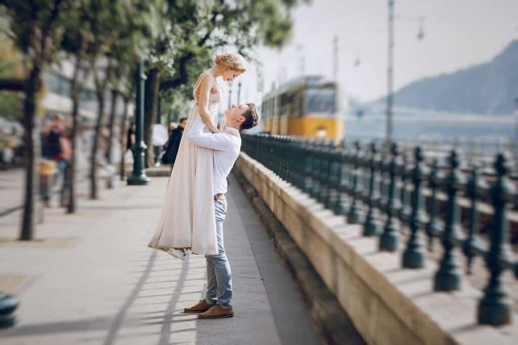 A man lifts a woman off the ground in an embrace on a walkway next to a wrought-iron fence, with a yellow tram in the background. They both appear happy and are dressed in light, casual clothing.