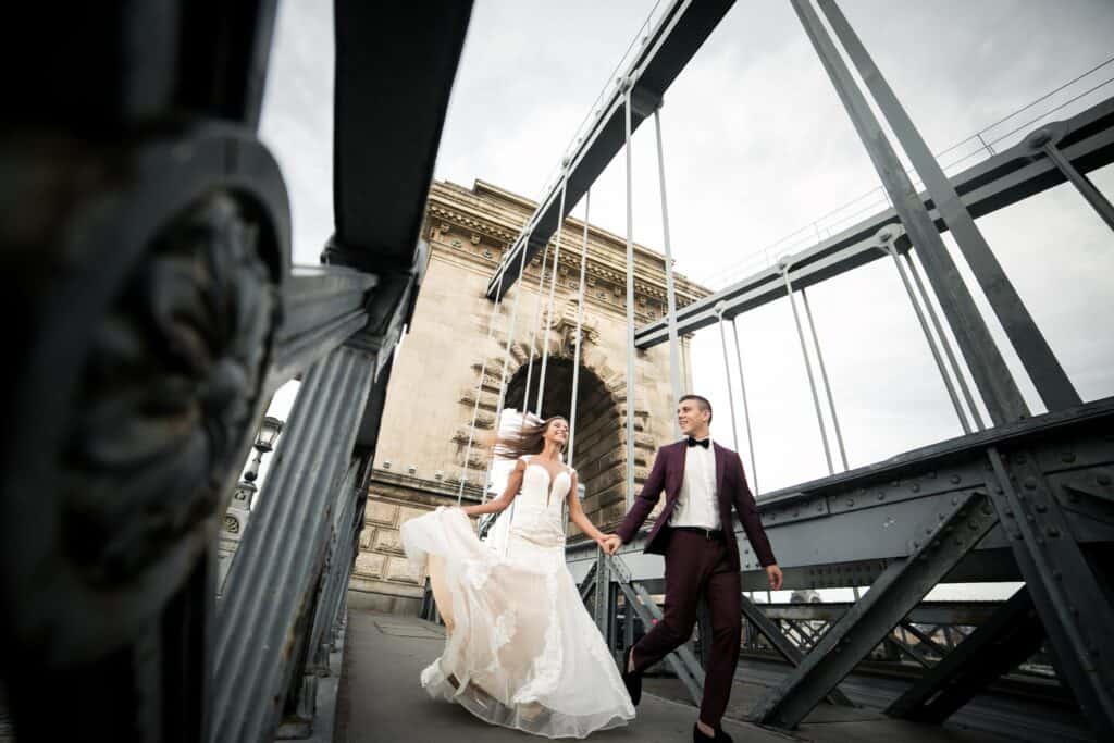 A couple in formal attire, with the woman in a wedding dress and the man in a suit, walk hand in hand on a steel bridge.