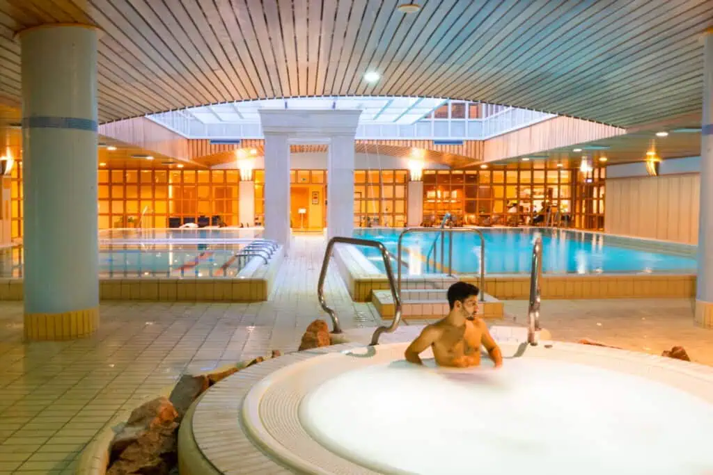 A man relaxes in a hot tub next to an indoor pool with wooden ceilings and walls.