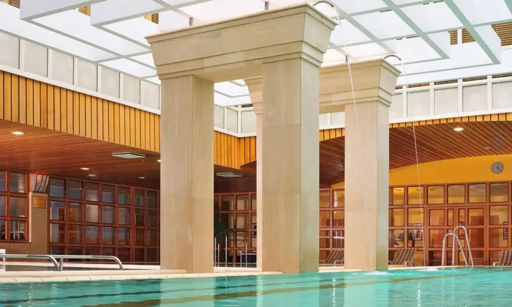 Indoor swimming pool area with large stone columns, wooden accents, a glass ceiling, and poolside seating.