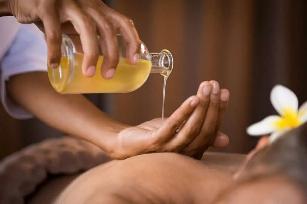 A person's hand pours massage oil from a glass bottle onto another hand over a person's back during a massage session.