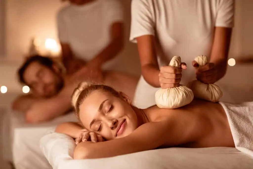 Two people receiving a massage in a dimly lit room; a woman in front smiles contentedly while a masseuse applies herb pouches to her back, and a man lies face down in the background receiving a massage.