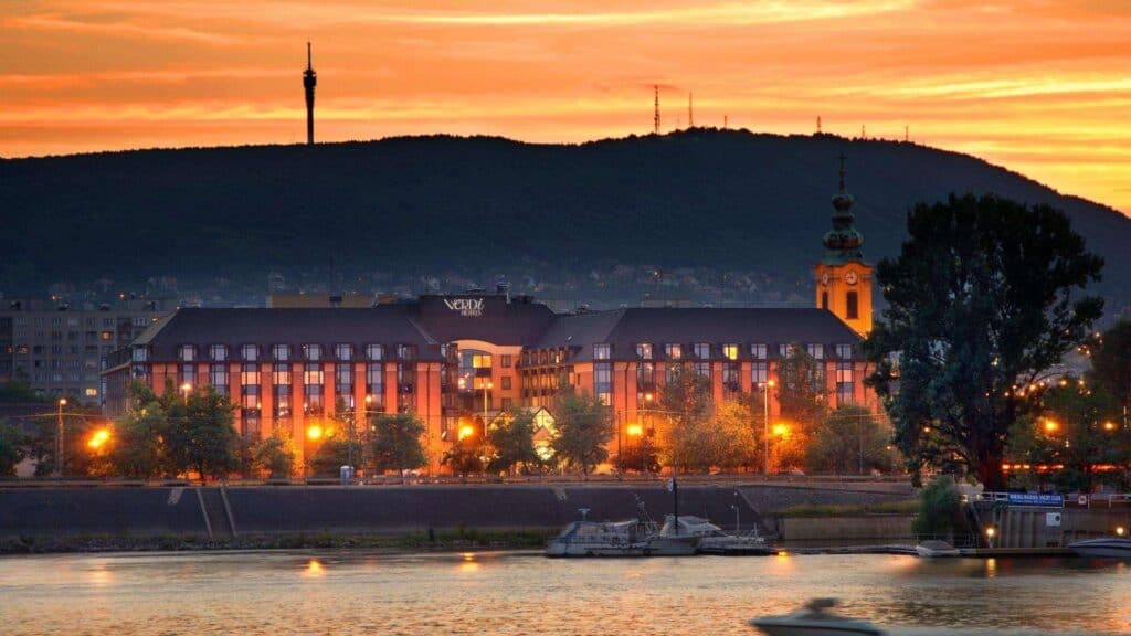 Riverside buildings with warm lights at sunset, hills and a tall tower in the background.
