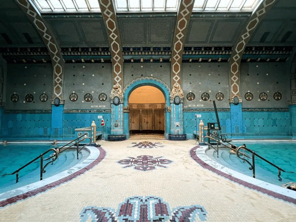 Ornate indoor swimming pool with blue and beige tiles, arched entrance, and decorative pillars. Skylights illuminate the space.