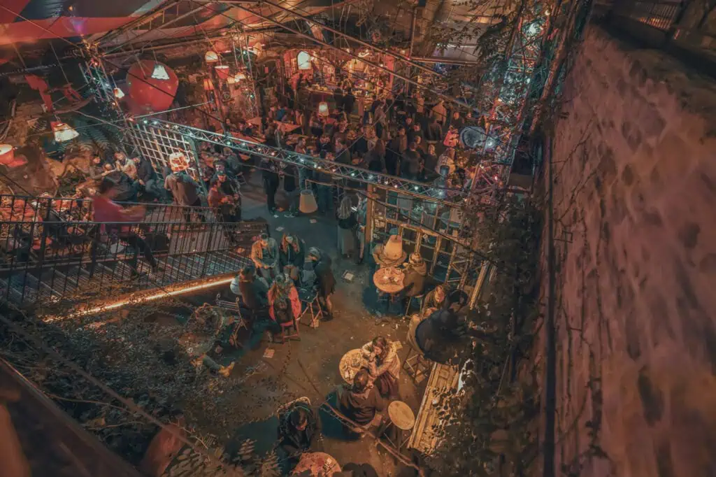 Aerial view of a crowded, dimly lit indoor venue with people sitting and standing around tables, colorful lighting, and eclectic decorations.