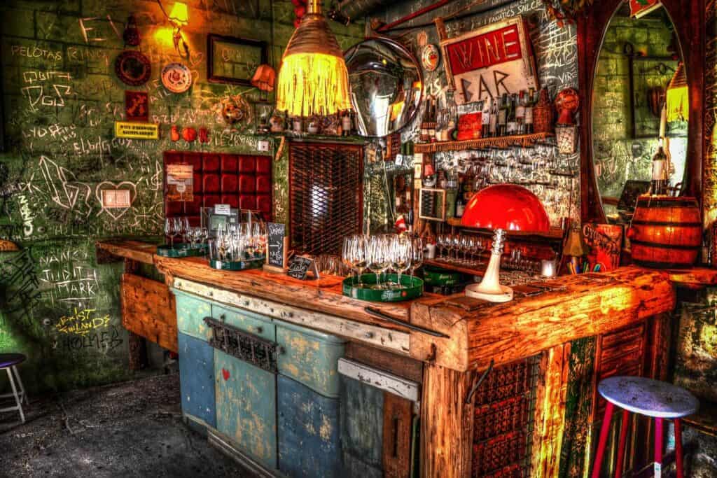 Rustic bar interior with wooden counter, vintage decor, hanging lights, and shelves filled with glasses and bottles. Walls are covered with chalk writing and quirky ornaments.