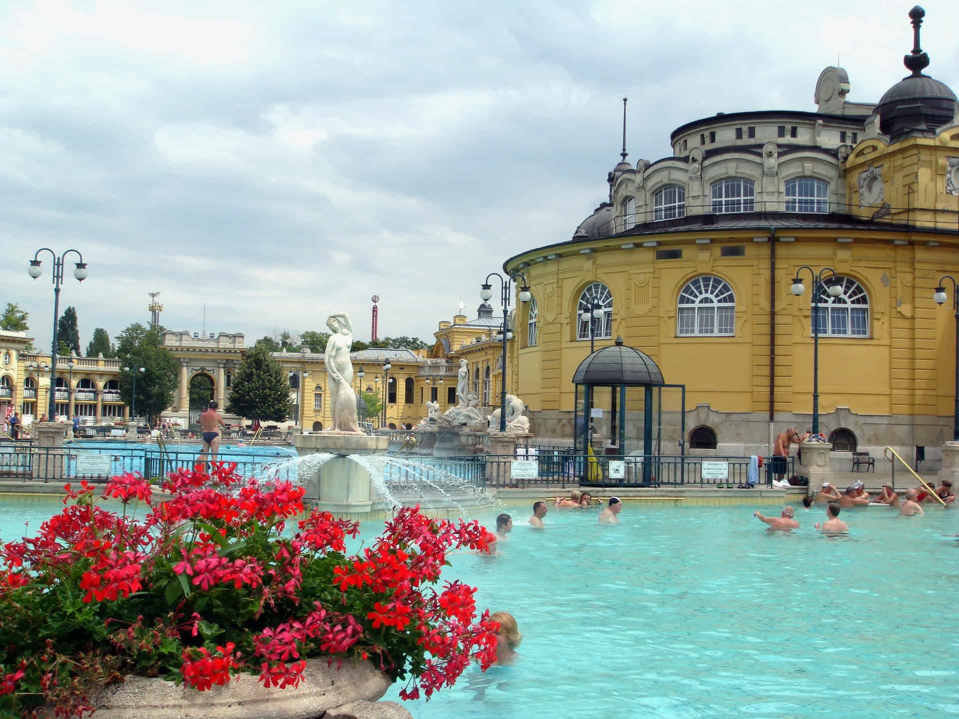 People are relaxing in a large outdoor thermal pool with a historic yellow building and red flowers nearby.