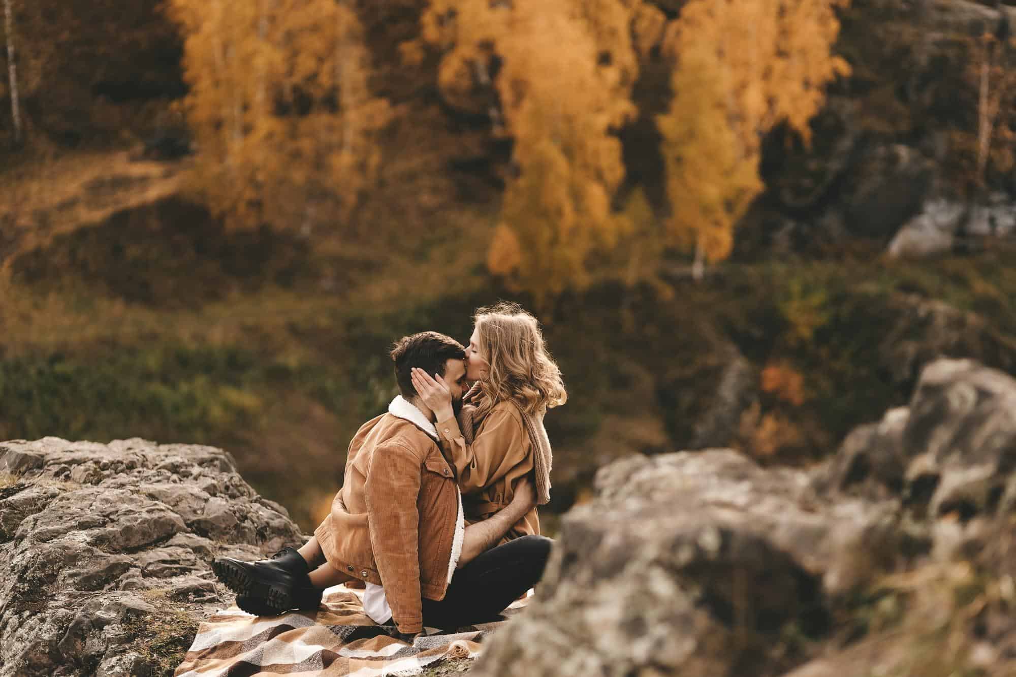 Two people sitting on a blanket share a kiss amidst a rocky landscape with vibrant autumn foliage in the background.