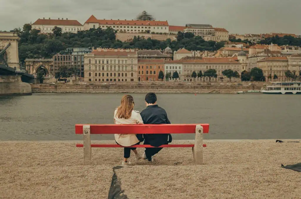 Two people sit on a red bench facing a river with a cityscape in the background.