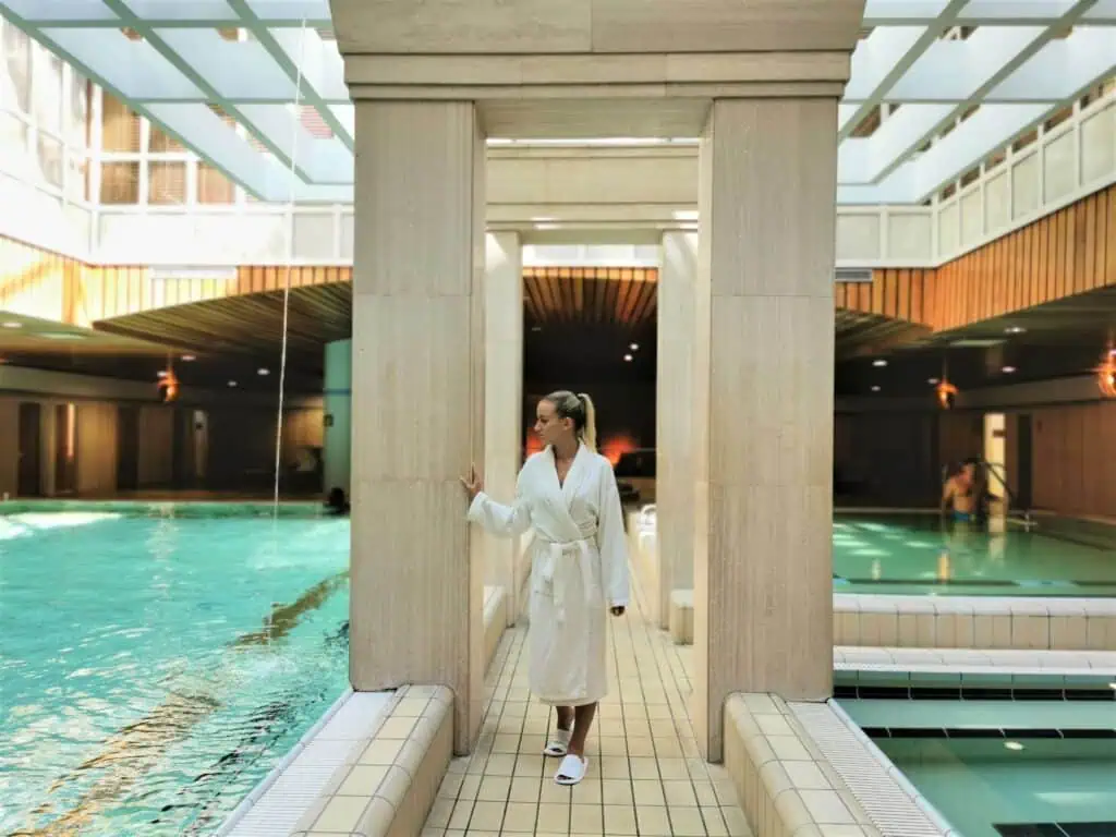 Person in a white robe and slippers standing by a swimming pool in an indoor spa setting with natural light filtering through a glass ceiling.