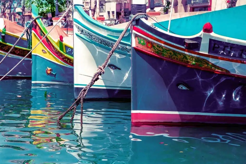 Colorful boats are moored in calm waters, with intricate patterns and painted eyes on their prows. The boats' reflections are visible in the water. Buildings and trees are seen in the background.