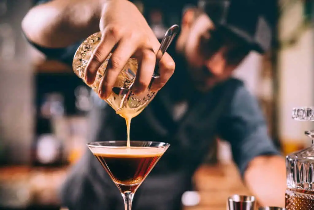 A bartender pours a cocktail with a creamy top into a martini glass using a cocktail strainer.