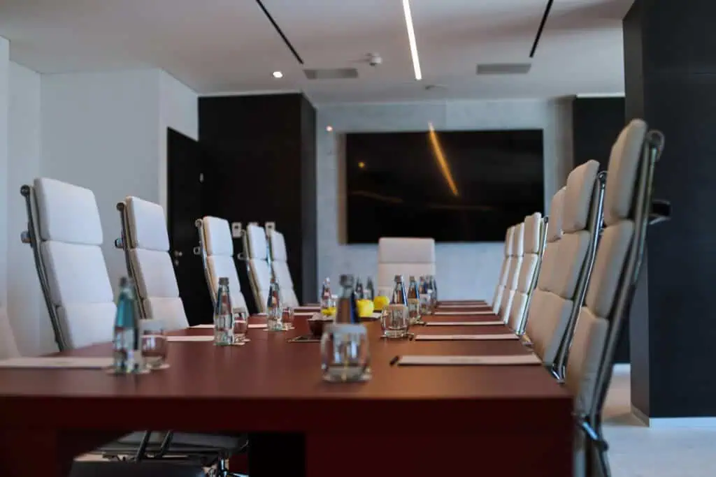 A modern conference room with a long table, white high-back chairs, bottled water, and notepads arranged on the table, and a large screen mounted on the wall in the background.