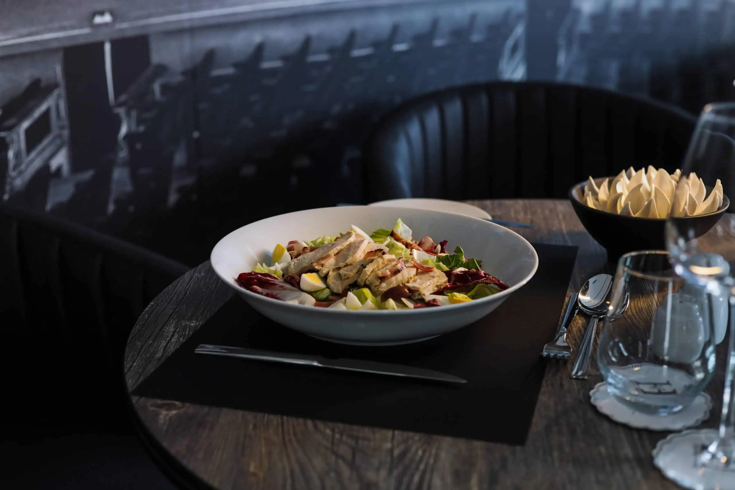 A salad with grilled chicken is served in a white bowl on a dark table, accompanied by a water glass, utensils, and a bowl of butter on the side.