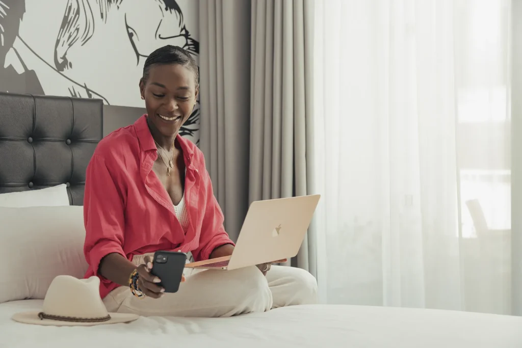 A person in a red shirt sits on a bed with a laptop on their lap and looks at a smartphone. A hat rests on the bed. Light filters through large curtains in the background.