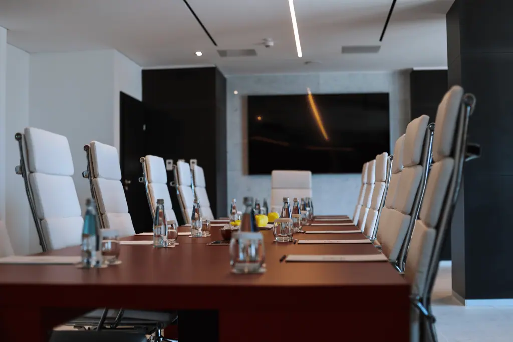 A modern conference room with a large table, high-back chairs, bottled water, glasses, and notepads, facing a large wall-mounted screen.