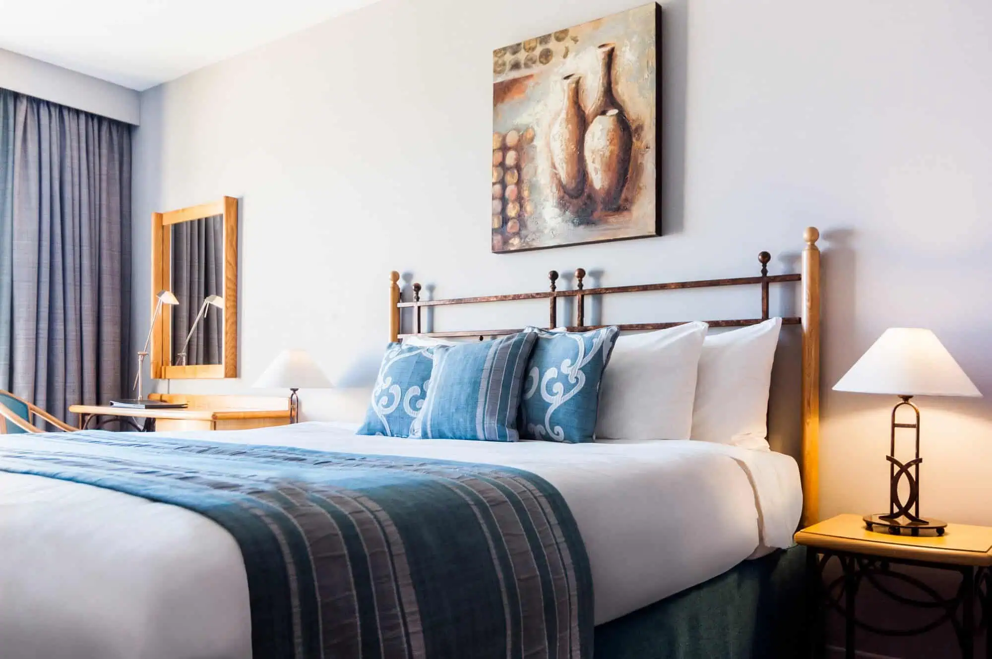 A neatly made bed with blue and white bedding in a well-lit room. A framed painting hangs above the headboard. There are two bedside tables with lamps and a wooden vanity with a chair.