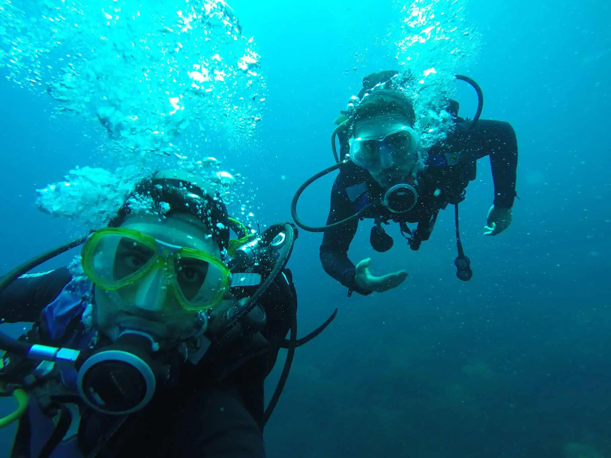 Two scuba divers underwater wearing diving masks and gear, surrounded by bubbles, in blue water.