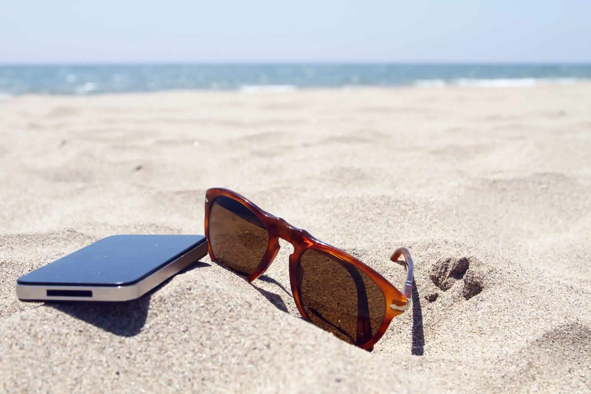 Sunglasses and a smartphone partially buried in the sand on a beach with the sea and sky in the background.