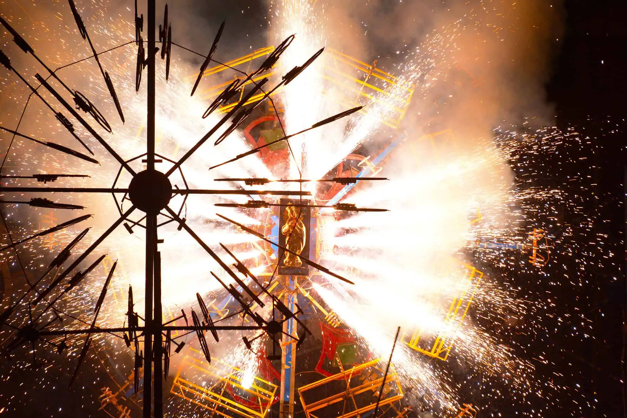 A structure with gears and spokes emits bright fireworks and sparks against a dark background.