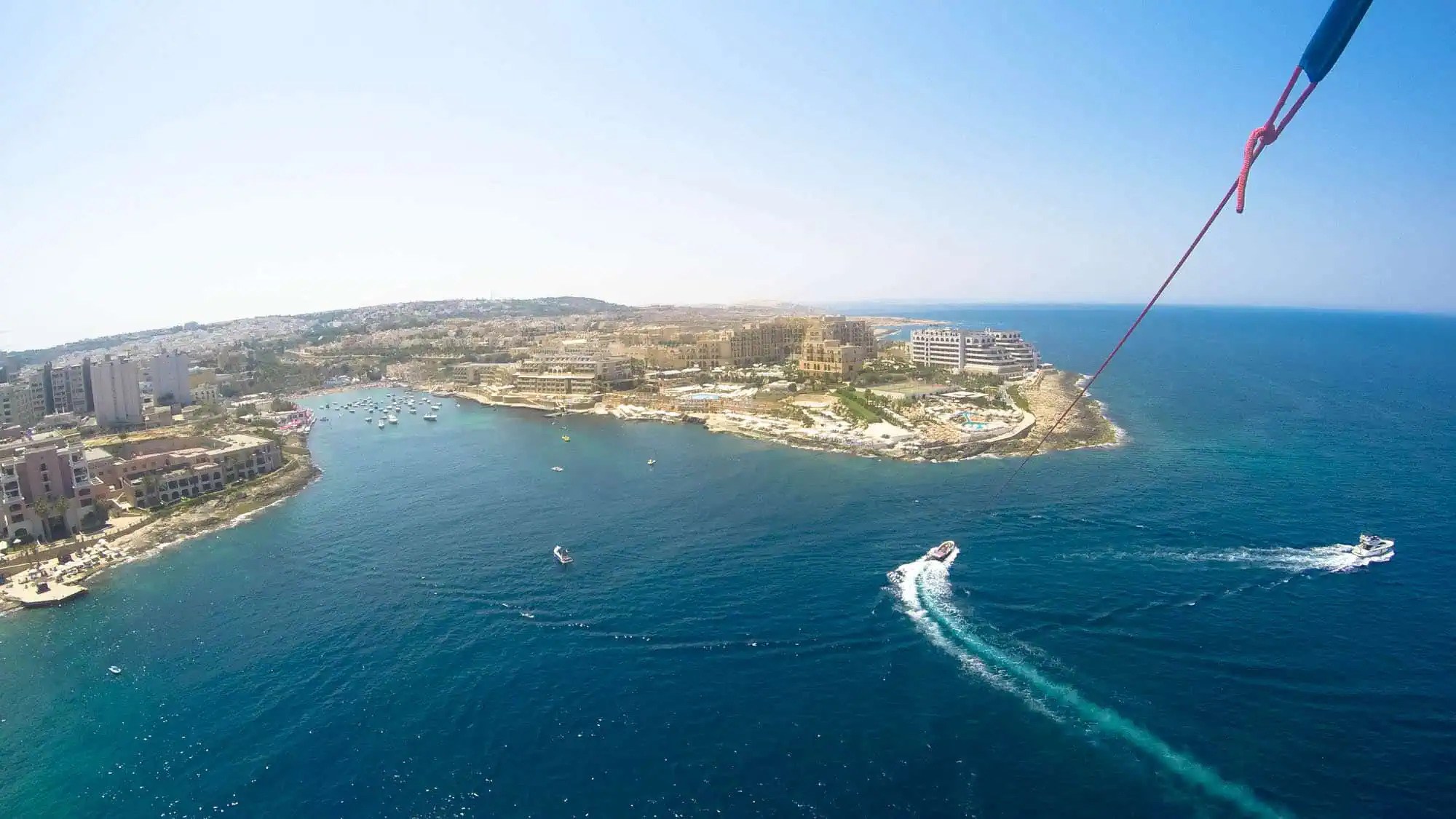 Aerial view of a coastal area with blue sea water, buildings along the shoreline, and a boat leaving a white trail in the water.