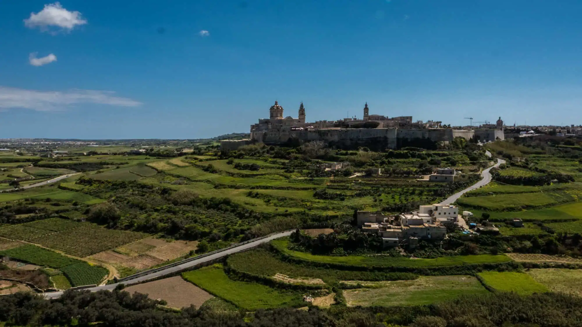 A historic city with church towers sits atop a hill, overlooking surrounding green fields and a winding road under a blue sky.