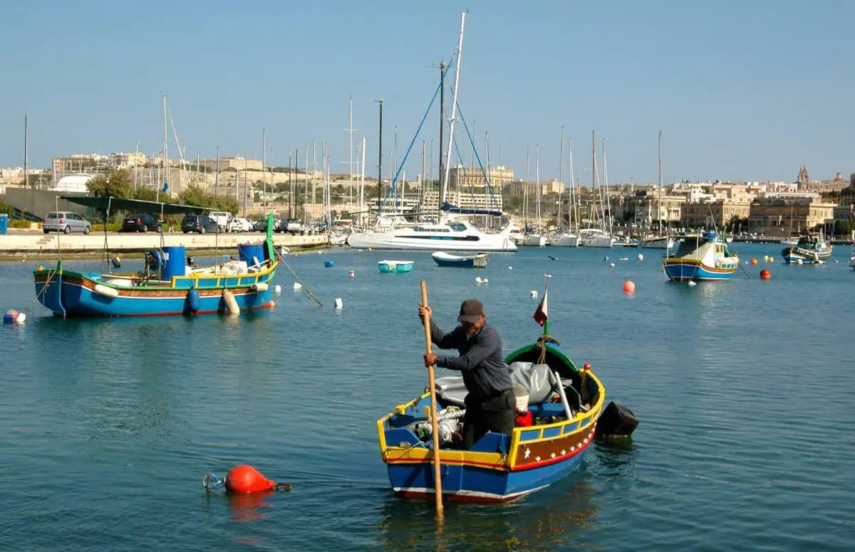 A person steers a small, colorful boat with a wooden pole in a harbor with other boats and yachts docked under a clear blue sky, embodying the charm of fishing in Malta.