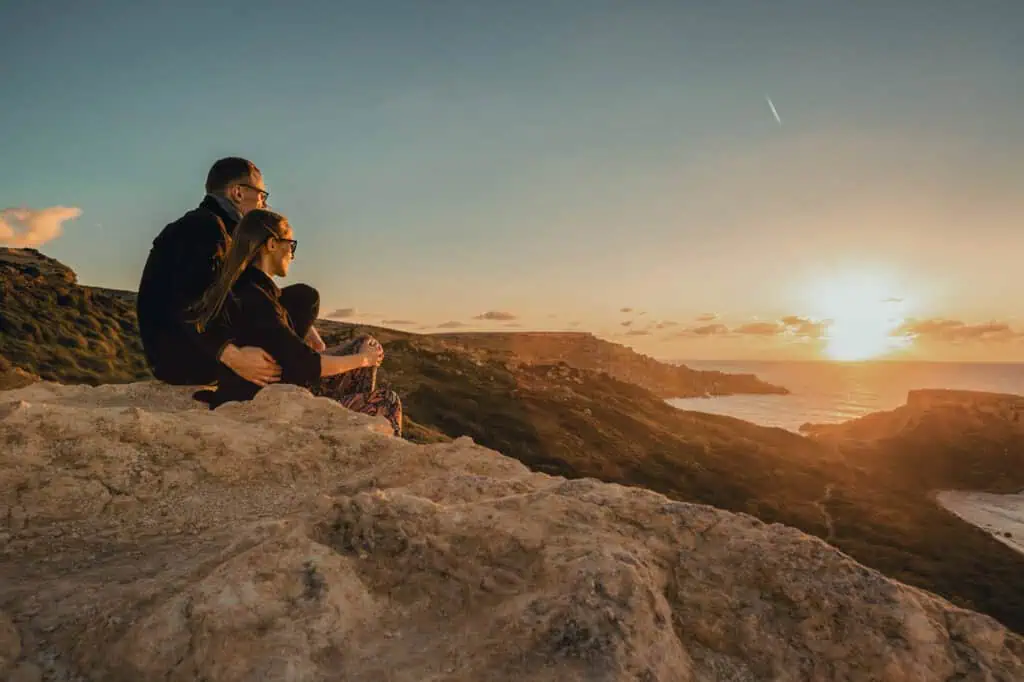 A couple sits on a rocky cliff, watching the ocean sunset, with a clear sky and distant clouds.