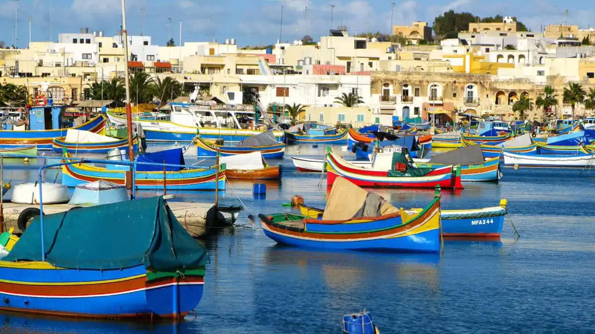 Colorful boats float in a harbor with a backdrop of a town featuring cream-colored buildings under a partly cloudy sky.